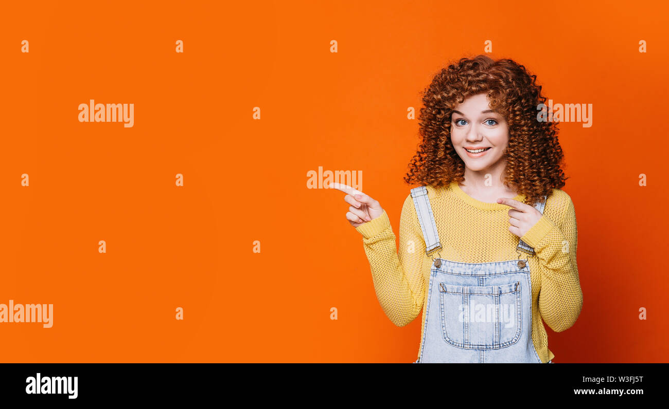 red-haired woman with curly hair advertising product, pointing hands on empty orange background Stock Photo