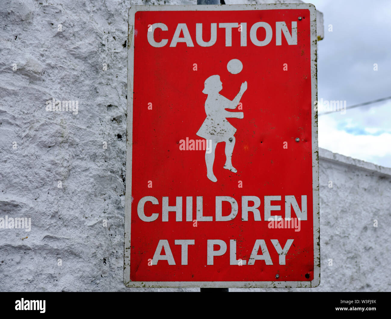 Sign Caution Children at play. Red background featuring girl playing ball, against a white textured wall Stock Photo