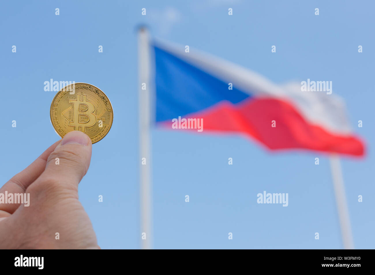 A man fingers are holding a big golden coin of bitcoin in front of Czech  Republic flag and blue sky.There is a big golden bitcoin coin on the Czech  Stock Photo -