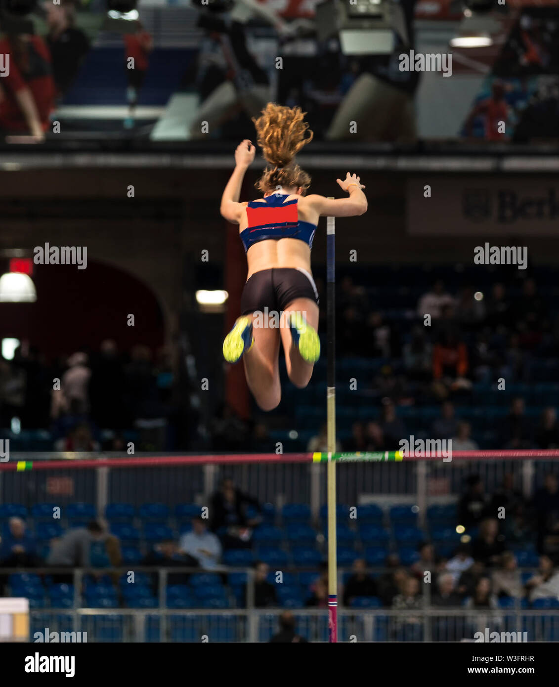 A high school female pole vaulter, athlete, is way over the bar in a competition in an indoor arena. Stock Photo