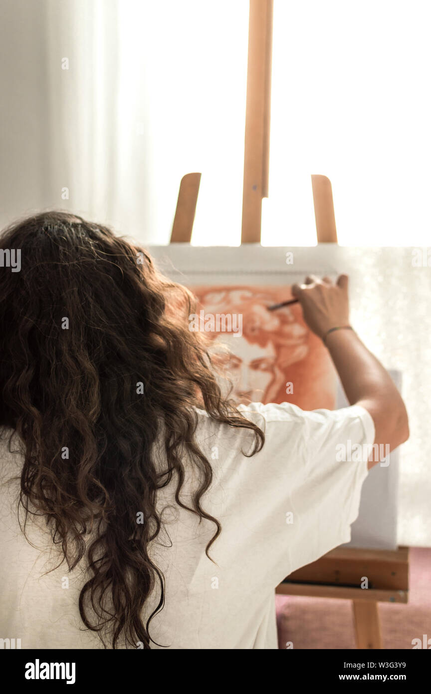 Woman with casual clothes painting with sanguine or red chalk. Drawing a greek portrait in front of a bright window with easel and orange pencil. Stock Photo
