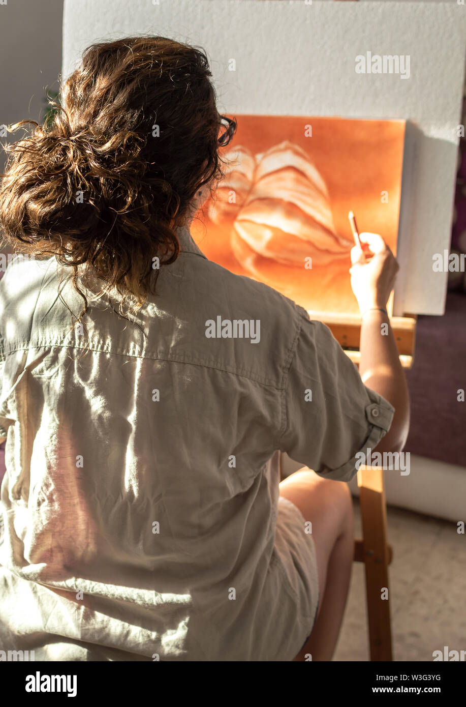 Woman with casual clothes painting with sanguine or red chalk. Drawing at home in front of a sunny window with easel. Stock Photo