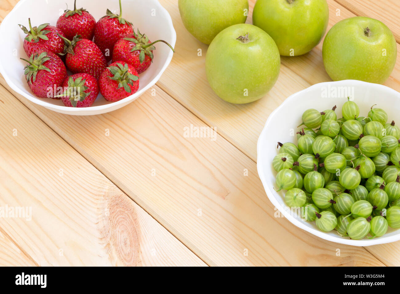 Green apples, gooseberry and red strawberries in white bowles on light wooden table. Background with fresh fruits and berries with copy space. Place f Stock Photo