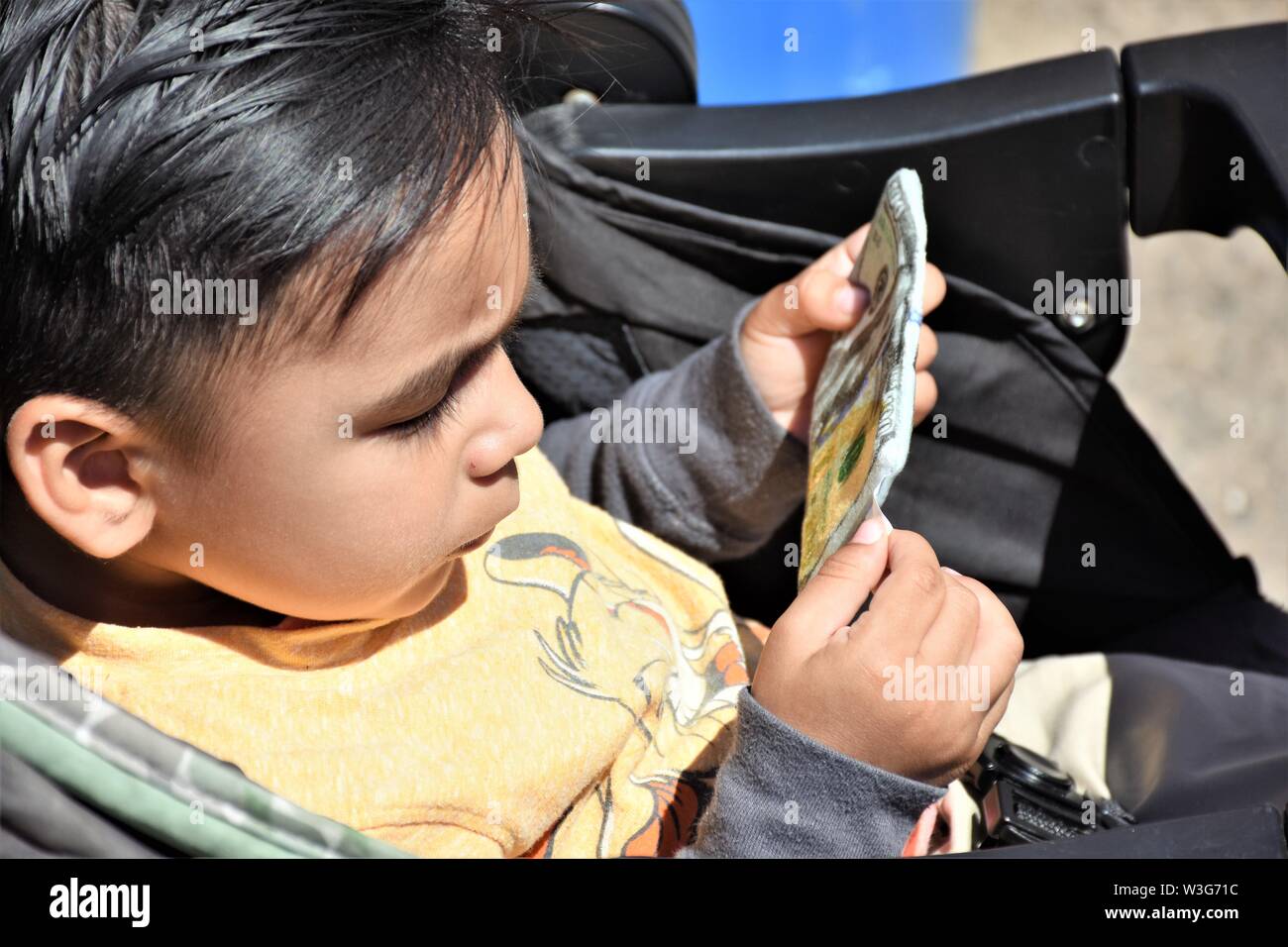 3 or 4 year old child playing with fake paper money hundred dollar bills in stroller frowning and watching real people at fair Stock Photo