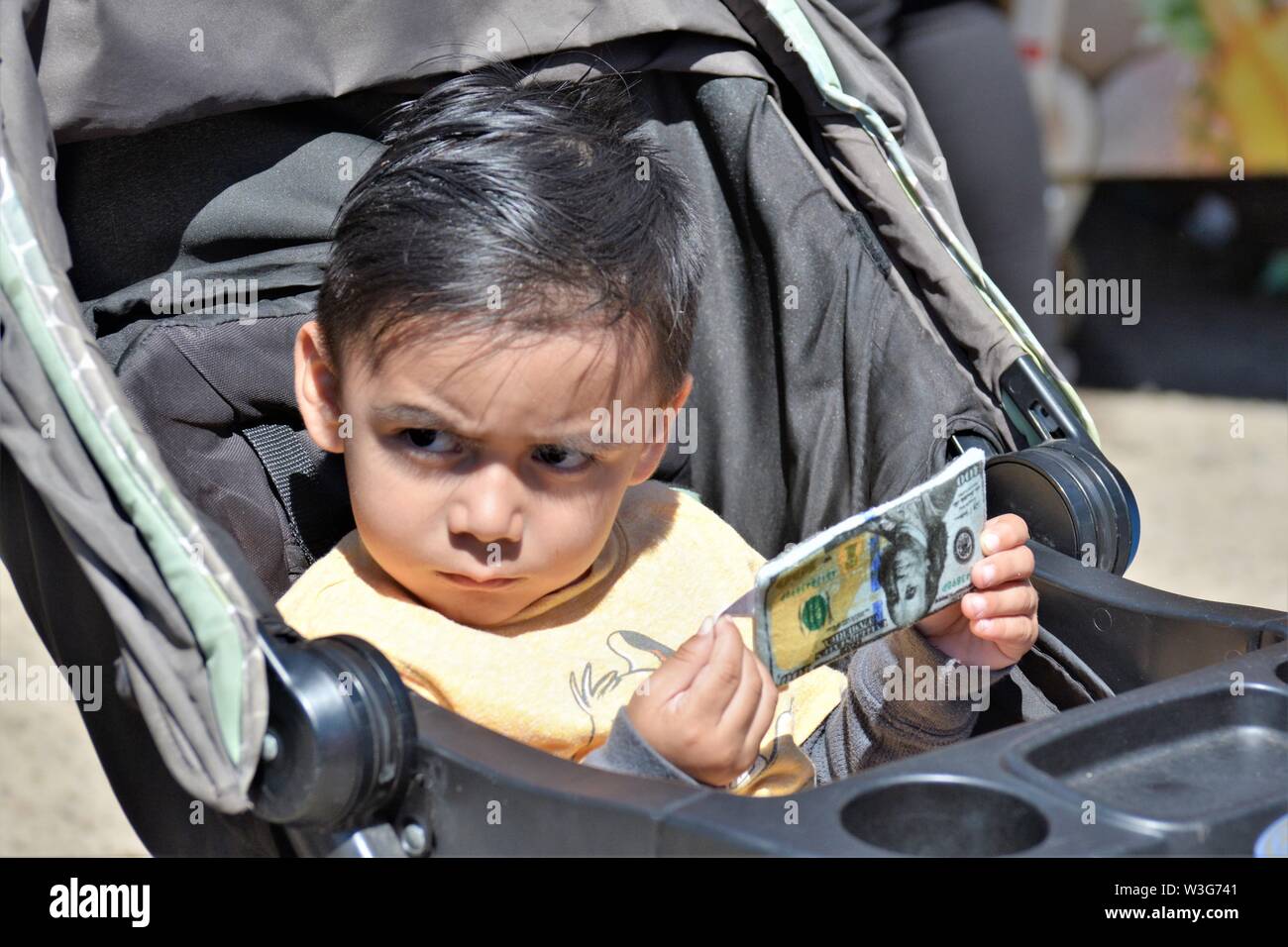 3 or 4 year old child playing with fake paper money hundred dollar bills in stroller frowning and watching real people at fair Stock Photo