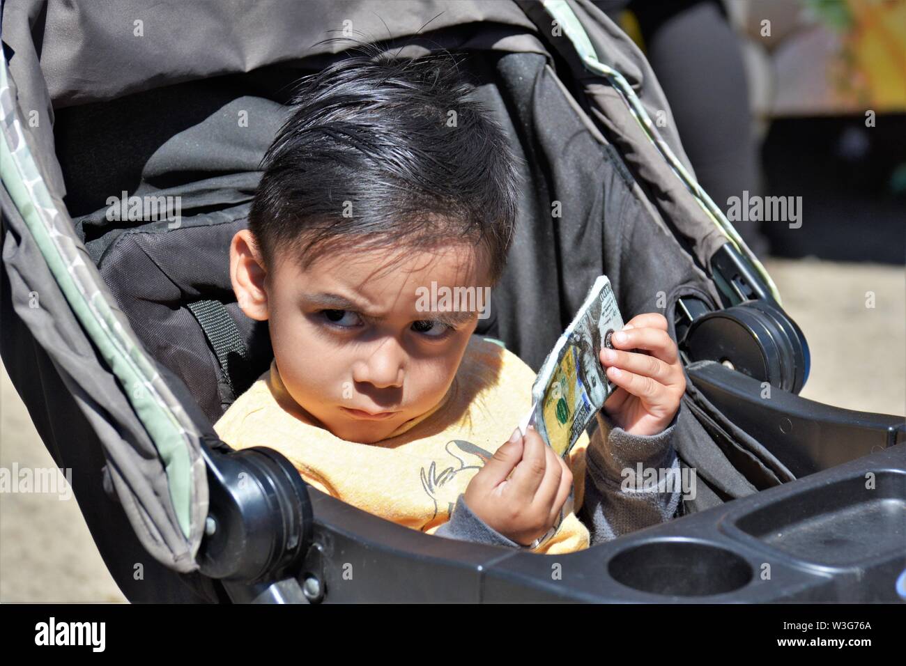 3 or 4 year old child playing with fake paper money hundred dollar bills in stroller frowning and watching real people at fair Stock Photo