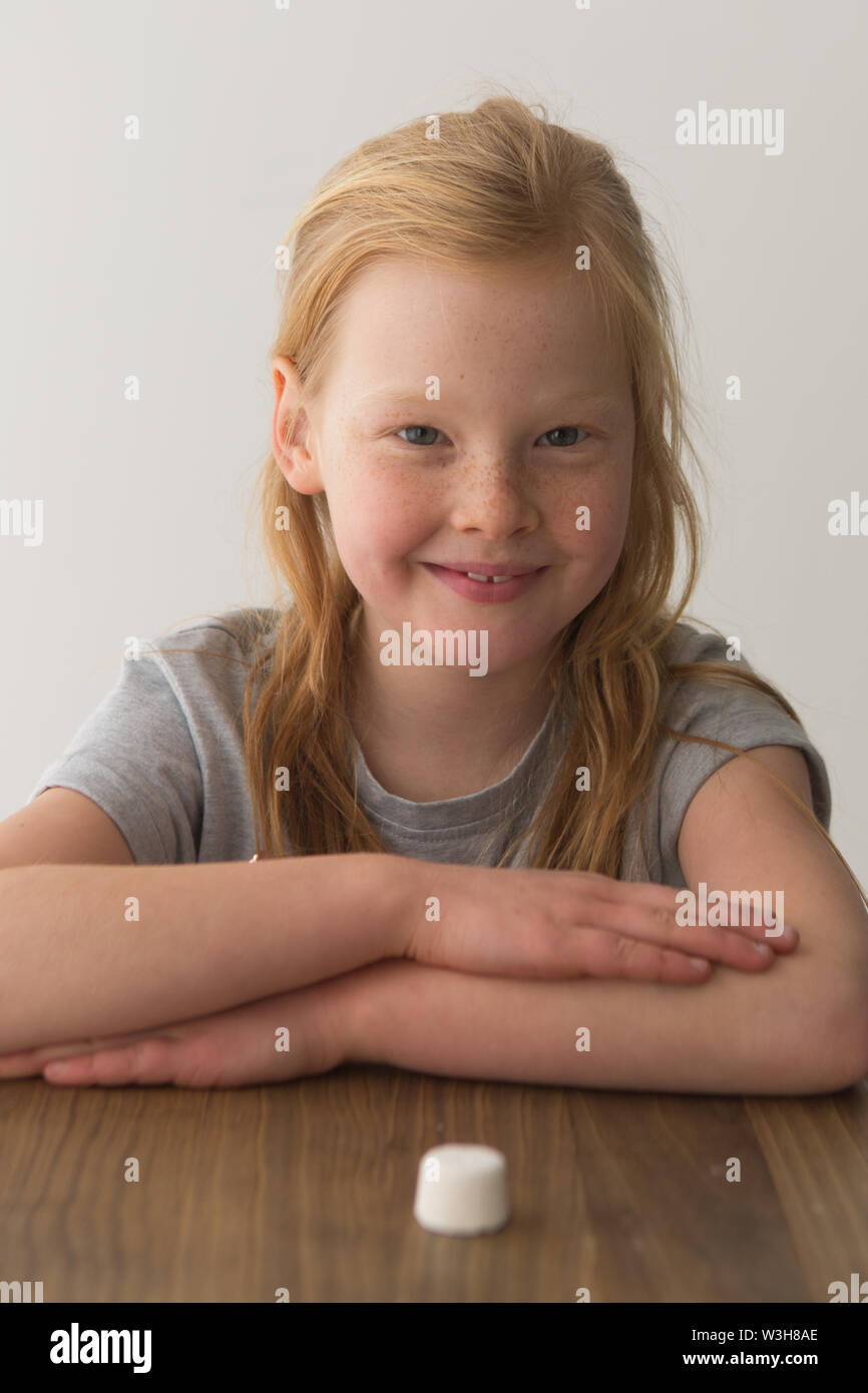 A young girl looks at a single marshmallow, attempting the marshmallow