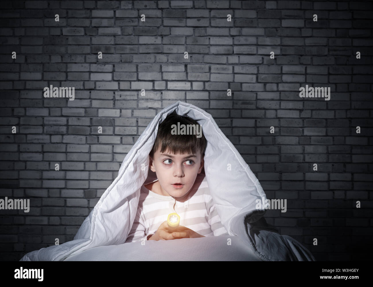 Frightened child with flashlight hiding under blanket. Scared kid lying