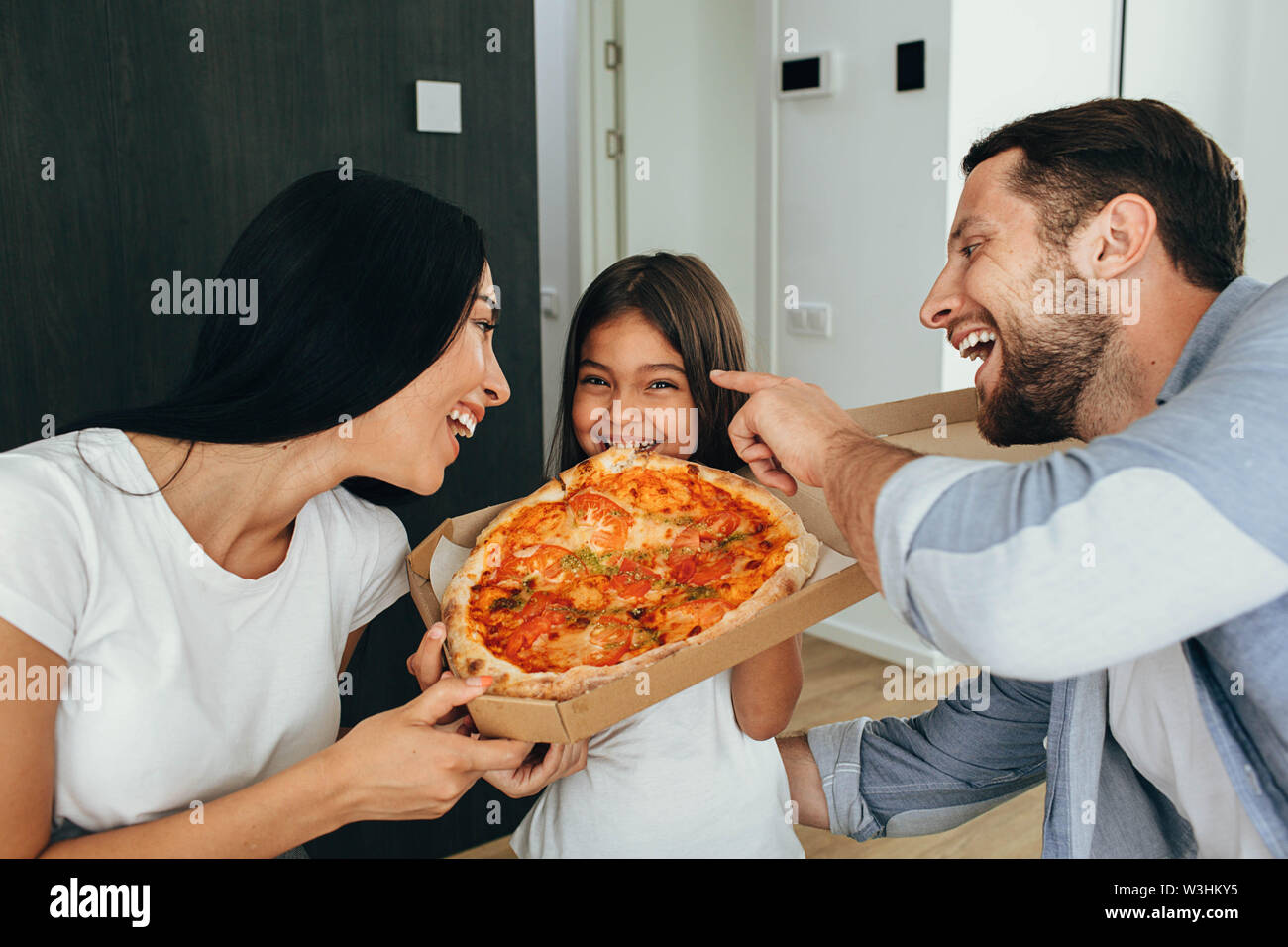 Eating tasty pizza with family. Funny little girl bitten piece of pizza, her parents laughing Stock Photo