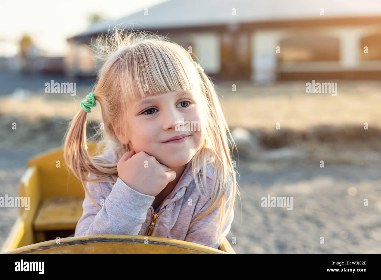 Adorable cute caucasian blond kid girl portrait sitting in wooden cart, looking aside and dreaming at farm or park during warm autumn evening Stock Photo