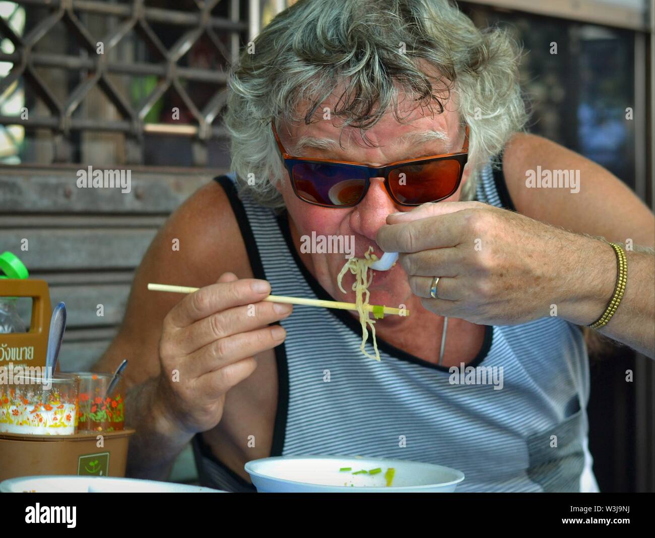 European tourist in Thailand eats spicy noodle soup with chopsticks and soup spoon. Stock Photo
