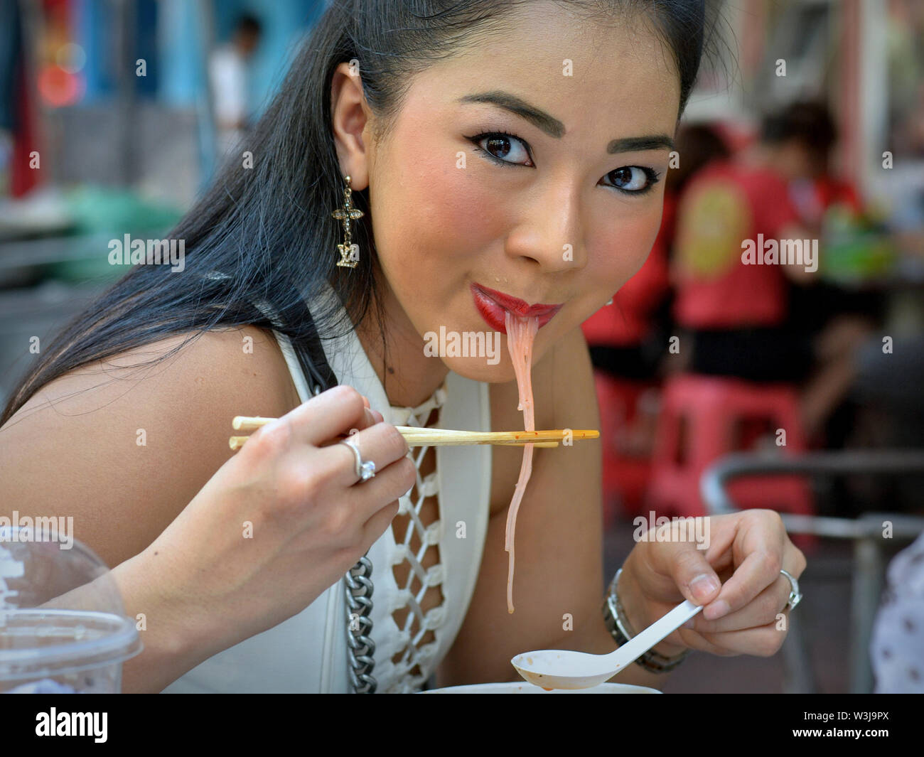 Glamorous Thai woman with beautiful eyes eats skillfully noodle soup with a pair of wooden chopsticks and a Chinese soup spoon. Stock Photo