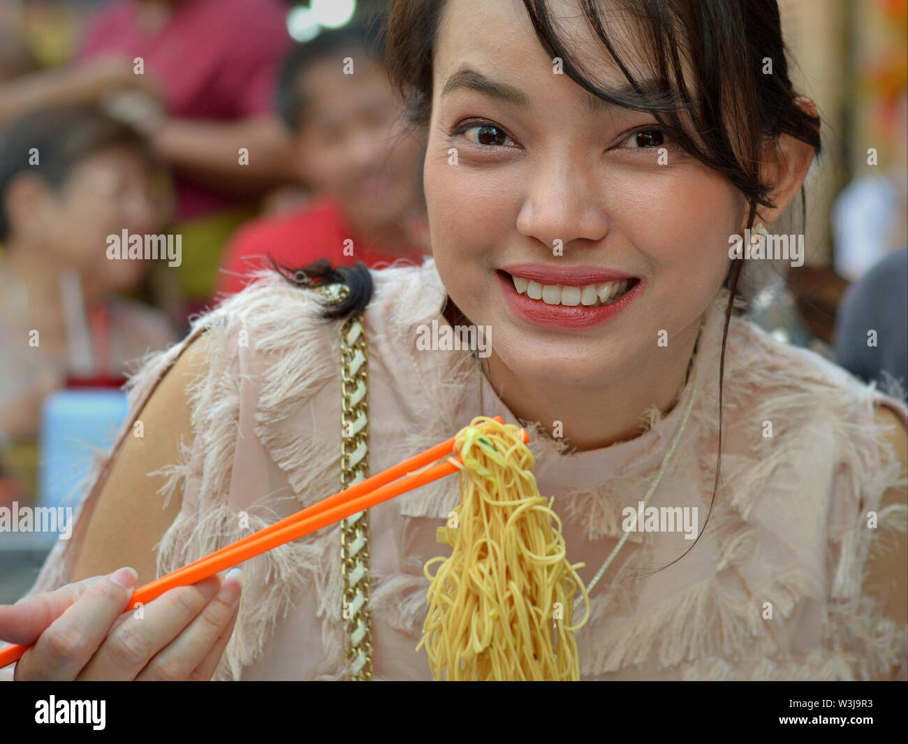 Glamorous Thai beauty eats skillfully dry yellow noodles with a pair of orange-colored plastic chopsticks and smiles for the camera. Stock Photo