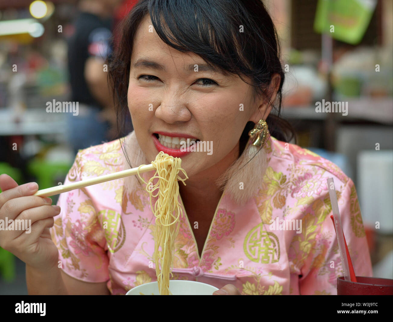 Dressed-up Thai Chinese woman with wrinkled nose eats yellow noodles with chopsticks during the Chinese New Year. Stock Photo