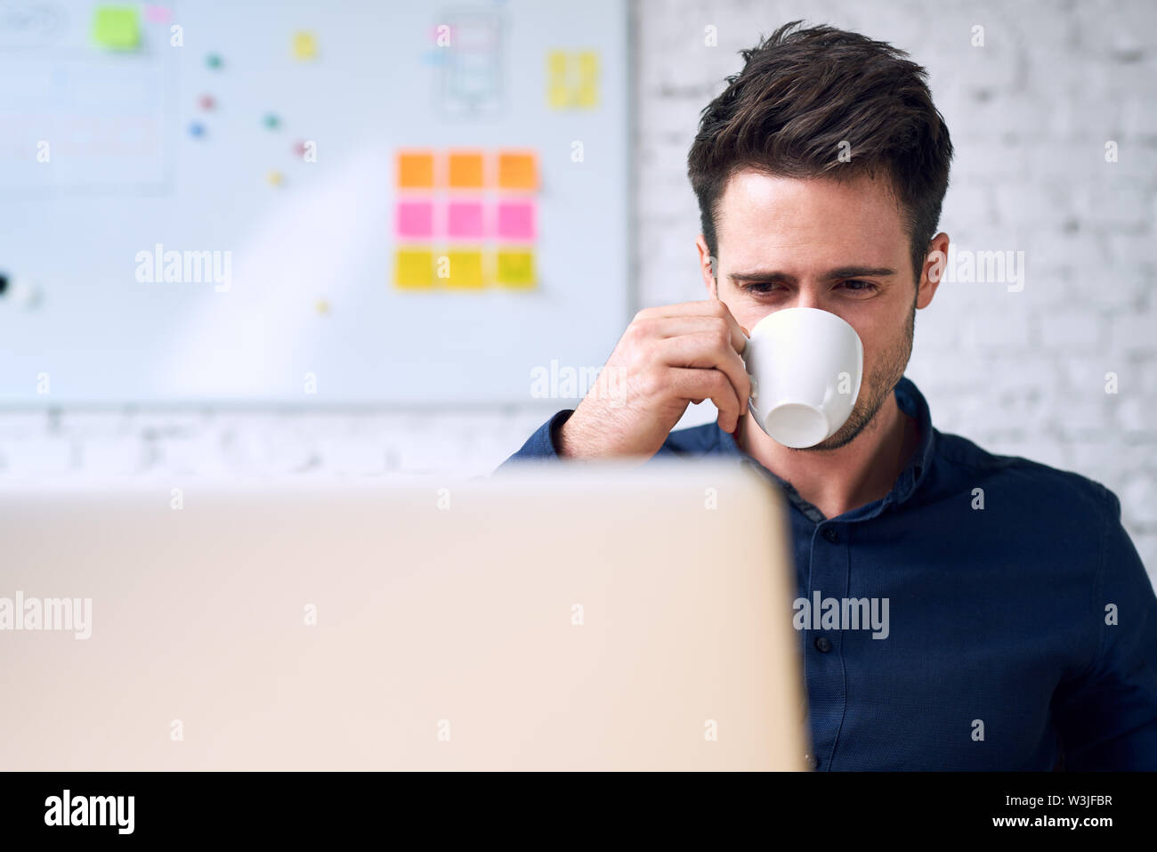 Young man, programmer drinking coffee looking on laptop screen. Stock Photo