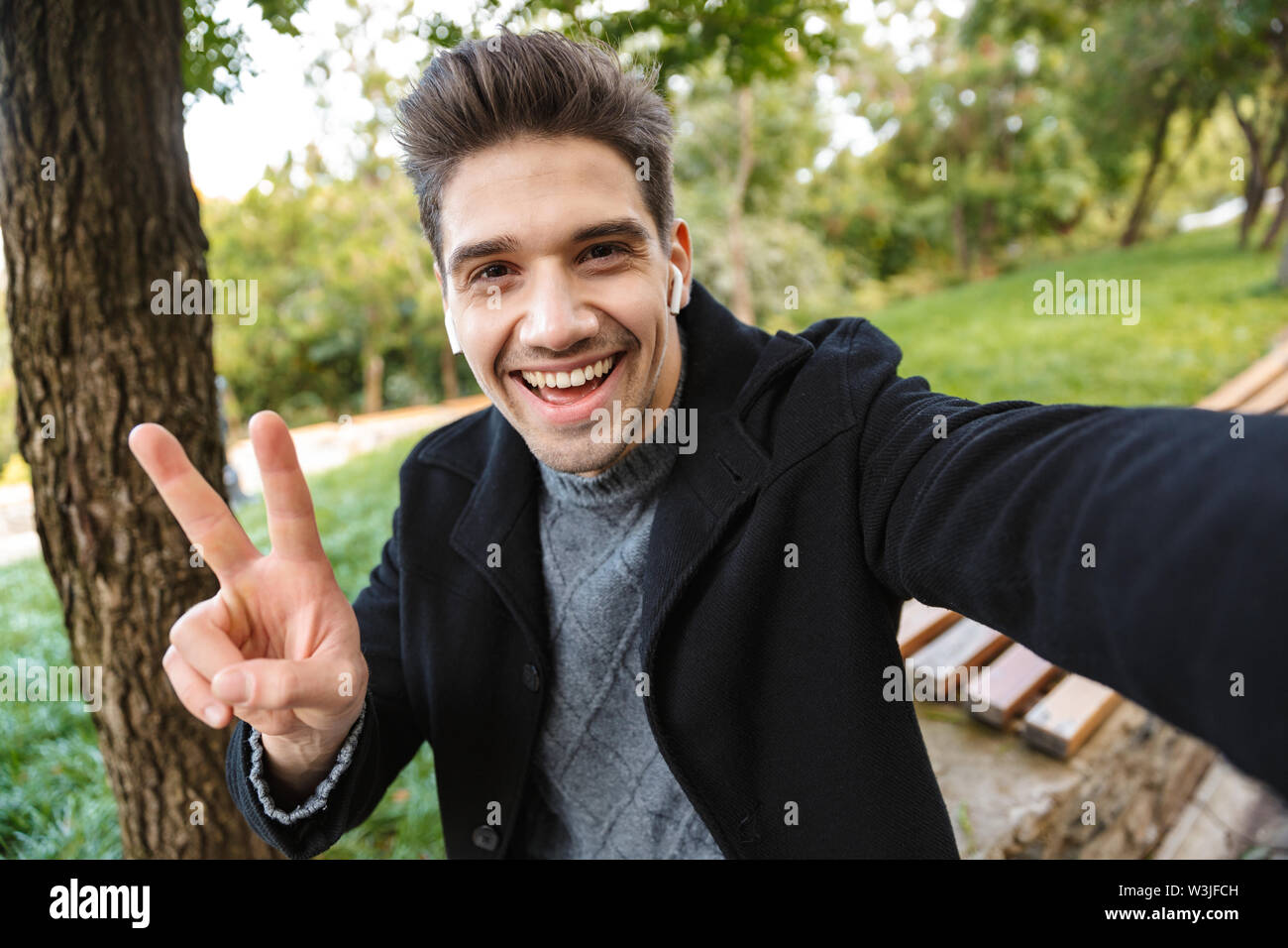 Image of cheerful young man in casual clothing walking outdoors in green park take a selfie by camera listening music with earphones showing peace ges Stock Photo