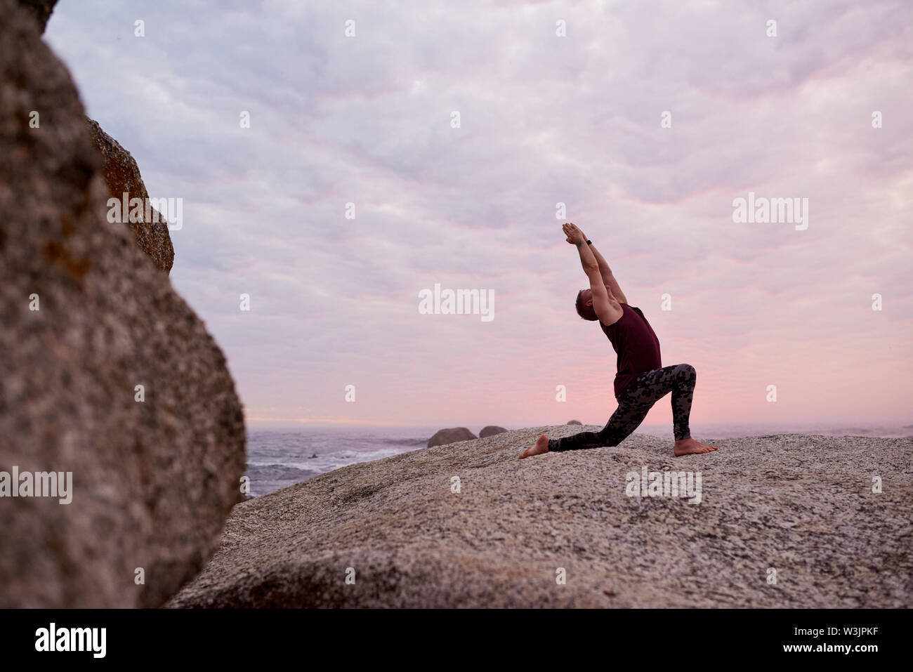 Man doing the cresent lunge pose on rocks at dusk Stock Photo