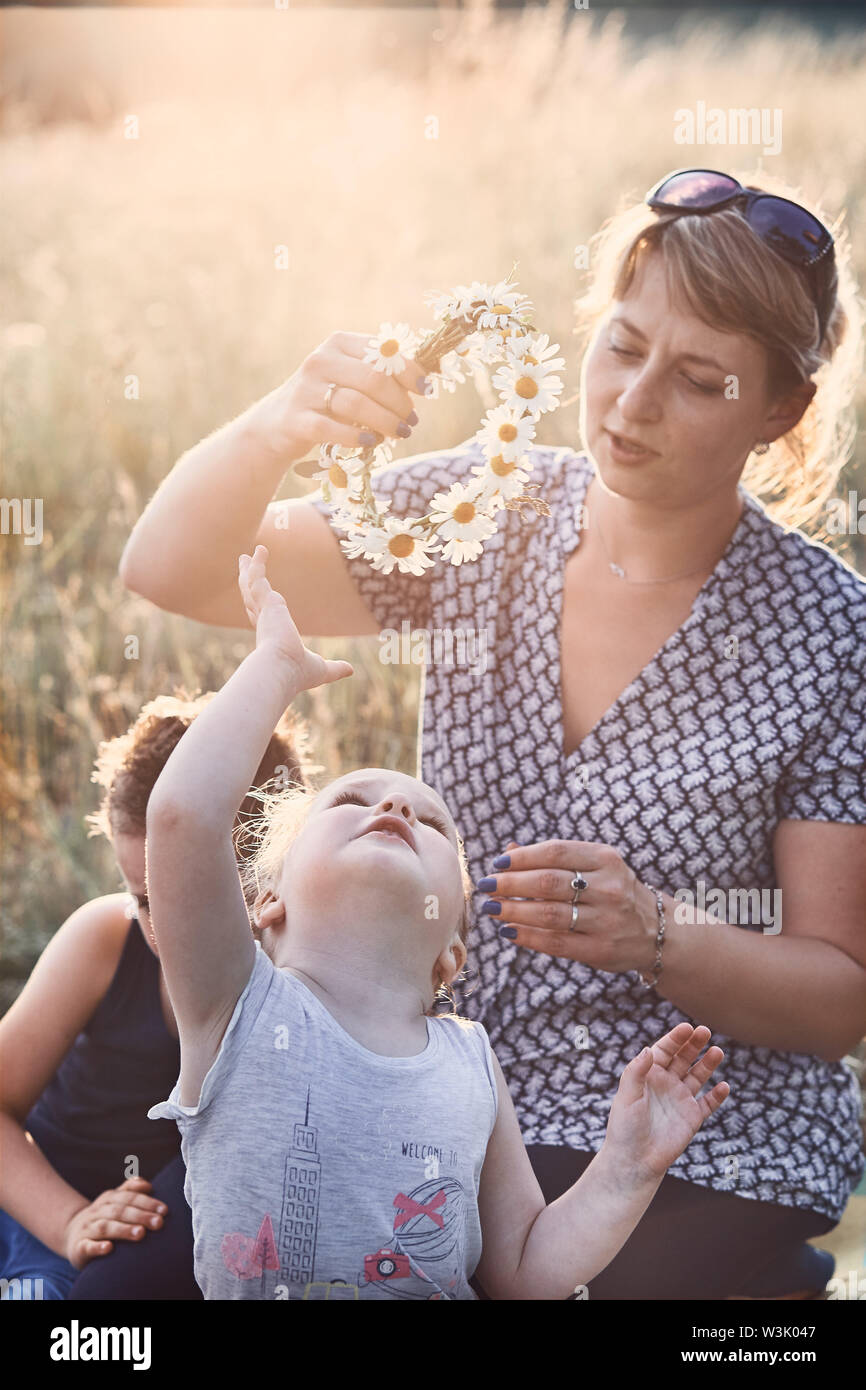 Mother putting a coronet of wild flowers on a head of little girl. Family spending time together on a meadow, close to nature. Parents and children pl Stock Photo