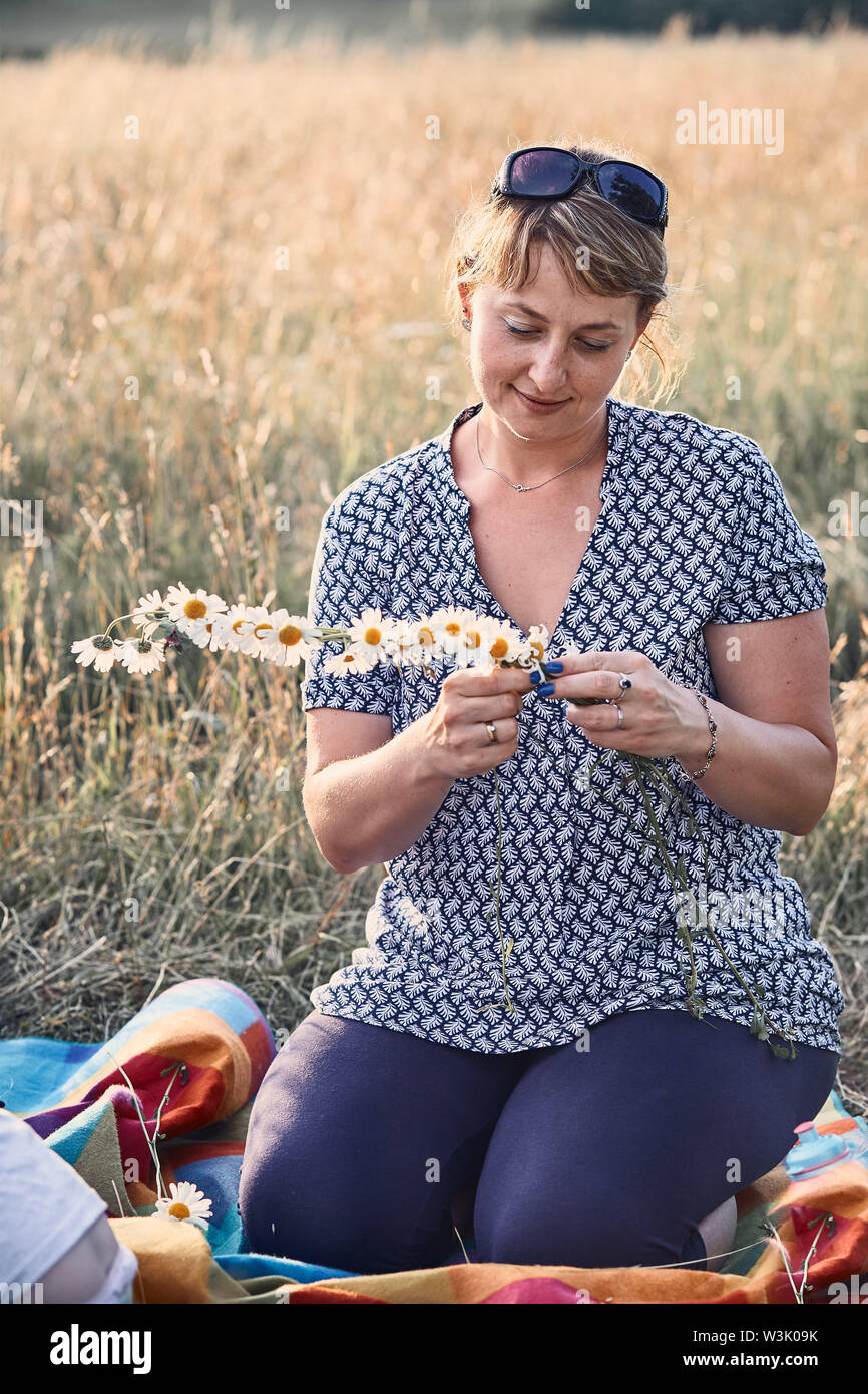 Smiling happy woman making coronet of wild flowers. Family spending time together on a meadow, close to nature. Candid people, real moments, authentic Stock Photo