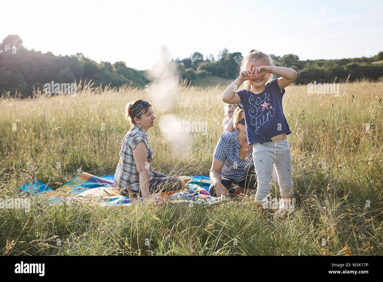 Little girl making a hand gesture for taking picture. Family spending time together on a meadow, close to nature. Parents and kids sitting on a blanke Stock Photo