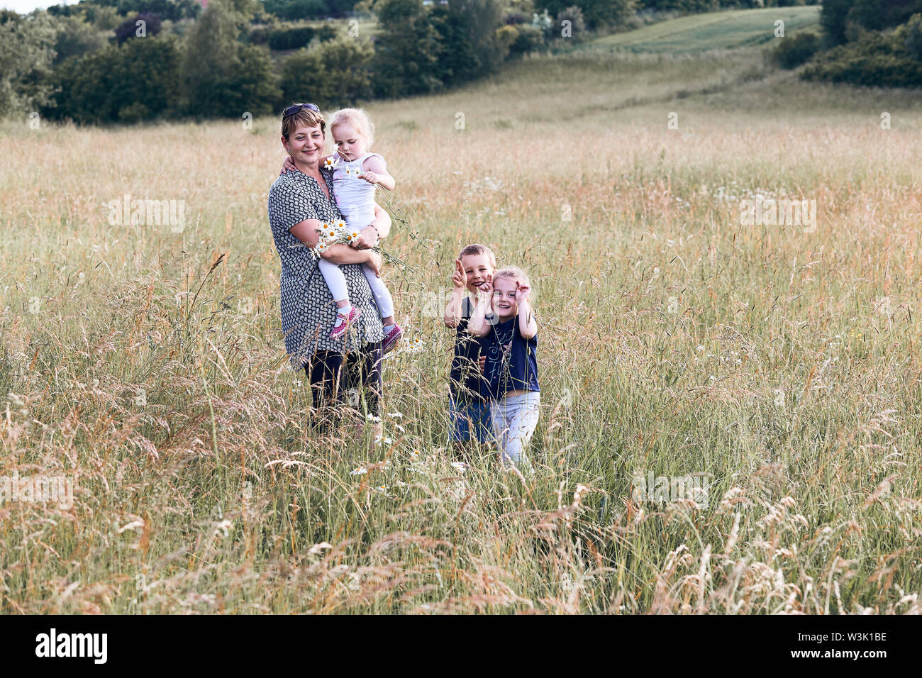 Mother spending time with children on a meadow, close to nature. Woman and kids standing in a grass. Happy kids making faces. Candid people, real mome Stock Photo