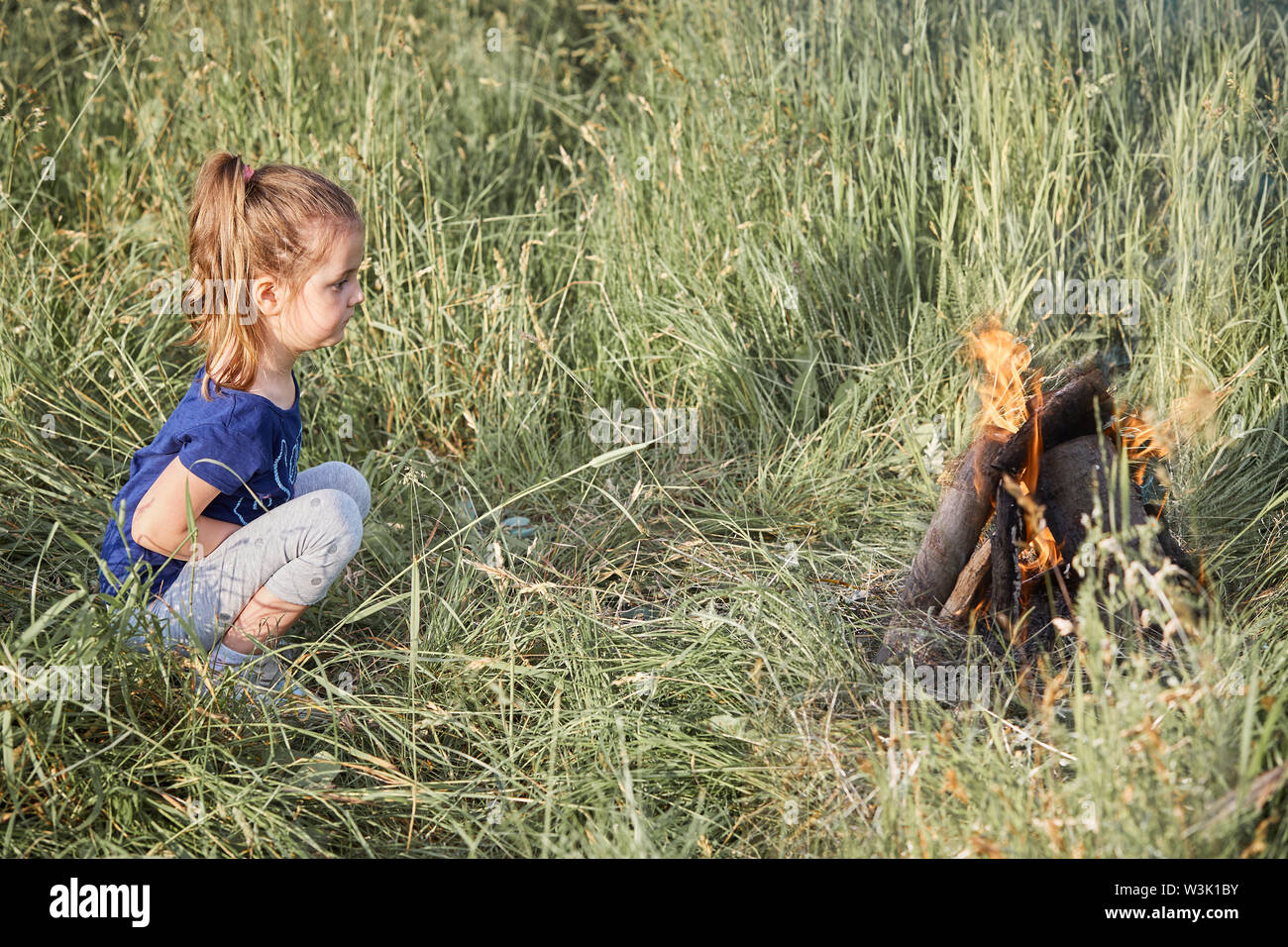 Little girl looking at campfire sitting in a grass. Candid people, real moments, authentic situations Stock Photo