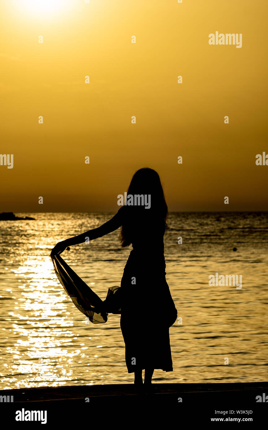 Beautiful female silhouette of unrecognizable slim tall woman with silk scarf at sunset in front of golden Ionian Sea water. Colorful dusk as seen from Ksamil, Albania, late spring Stock Photo