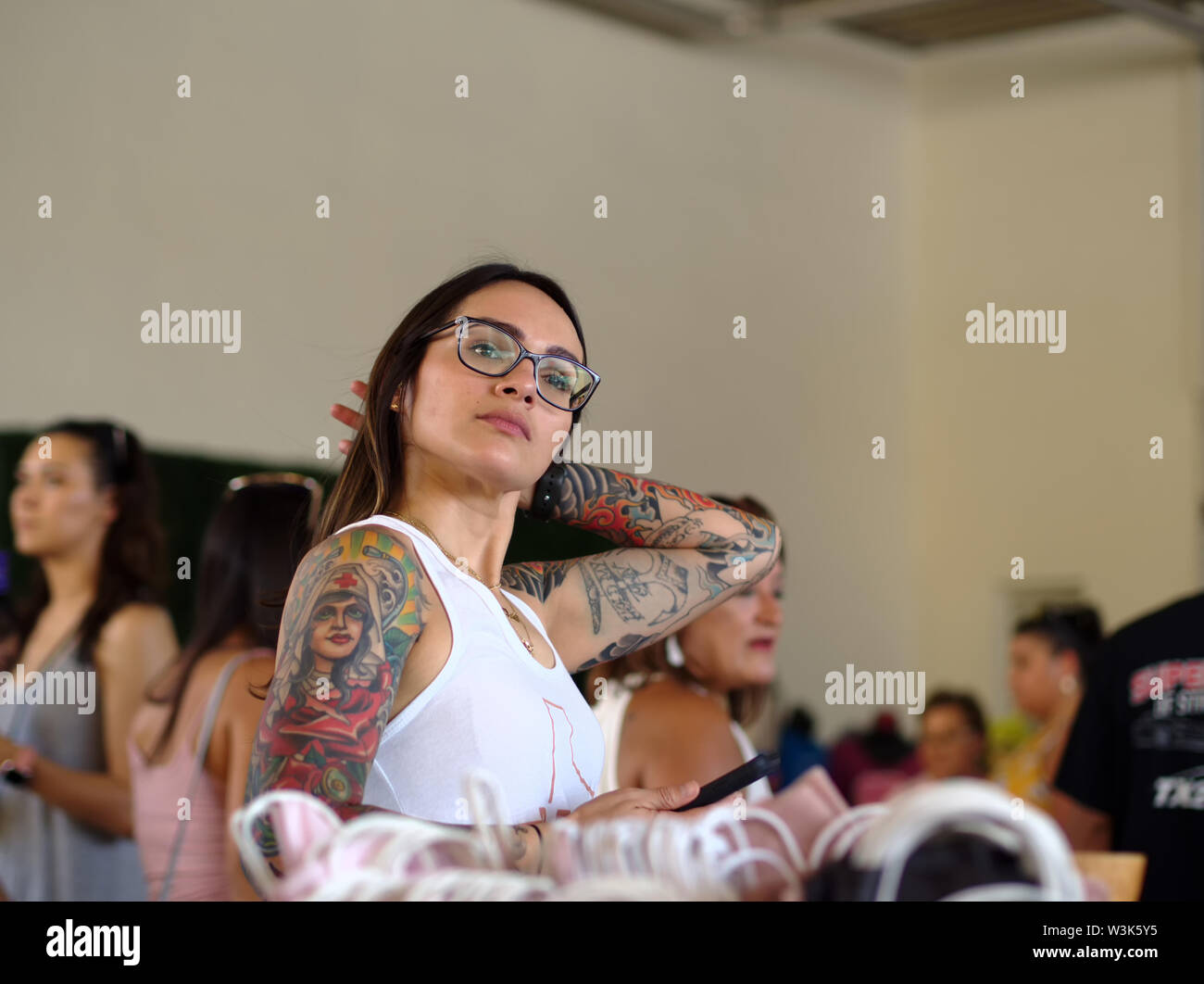 An attractive, heavily tattooed, young brunette in white tank top and glasses pulls her hair back. 'Loca for Local' event Corpus Christi, Texas USA. Stock Photo