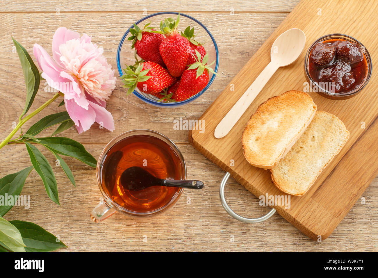 Traditional homemade strawberry jam in a glass bowl, fresh berries, toasts on cutting board with spoon, cup of tea and peony flower on wooden desk. To Stock Photo