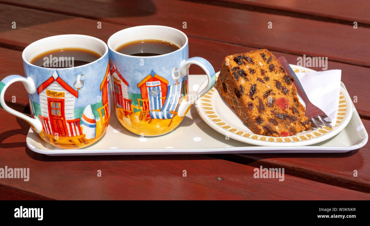Devon, England, UK. July 2019. Fruit cake on a plate with two decorated cups of black coffe on a tray Stock Photo