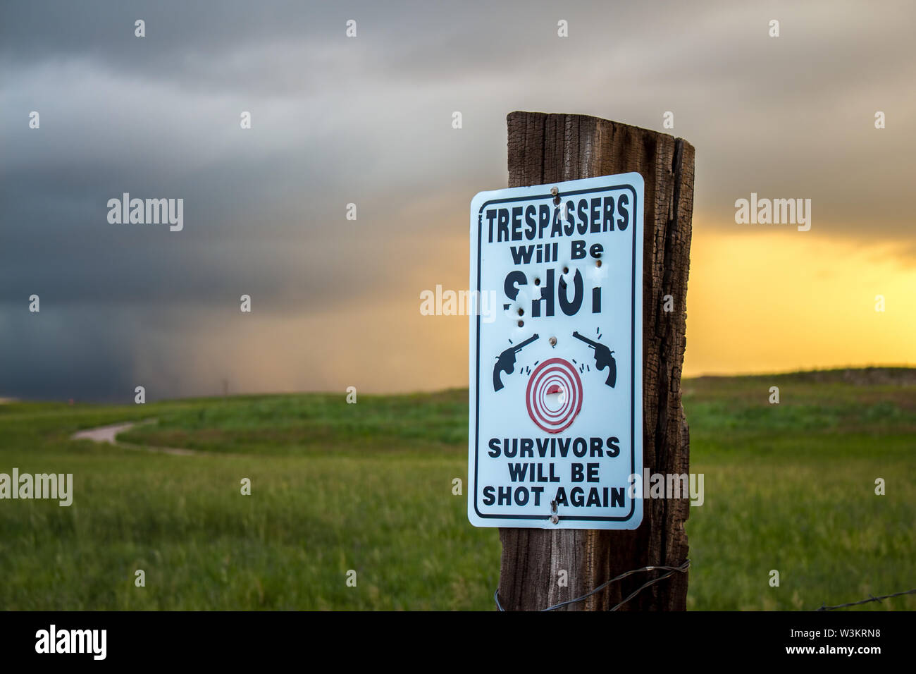 A no trespassing sign riddled with bullet holes reads: Trespassers will be shot. Survivors will be shot again. Stock Photo