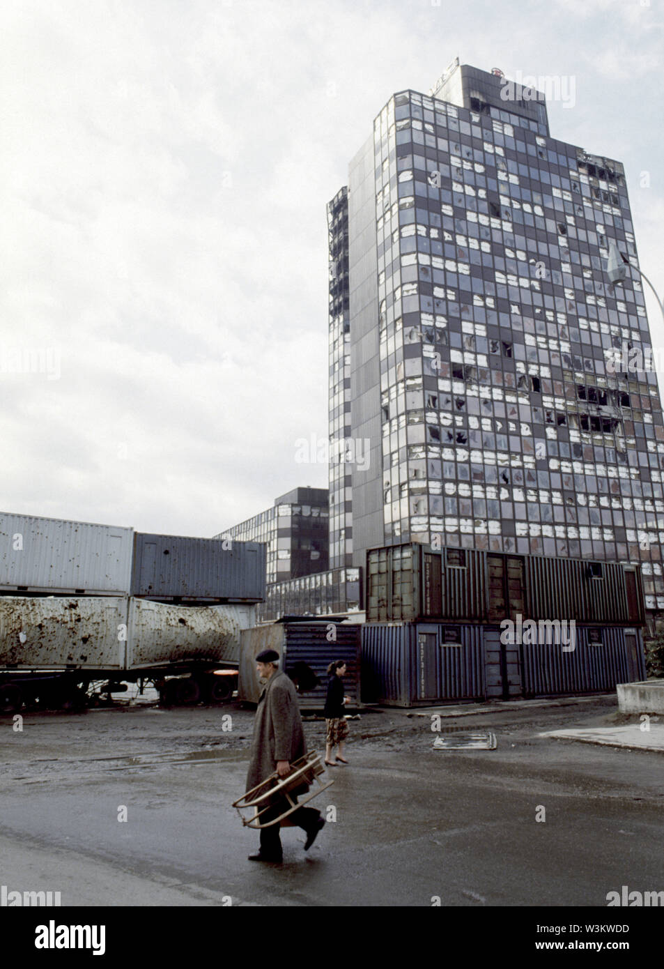 11th April 1993 During the Siege of Sarajevo: in the shadow of the Energoinvest buiding, local people walk past shrapnel-ridden containers that act as a sniper screen at the junction of Bratstva i jedinstva and Krusevacka (renamed Hamdije Cemerlica and Put zivota after the war). Stock Photo