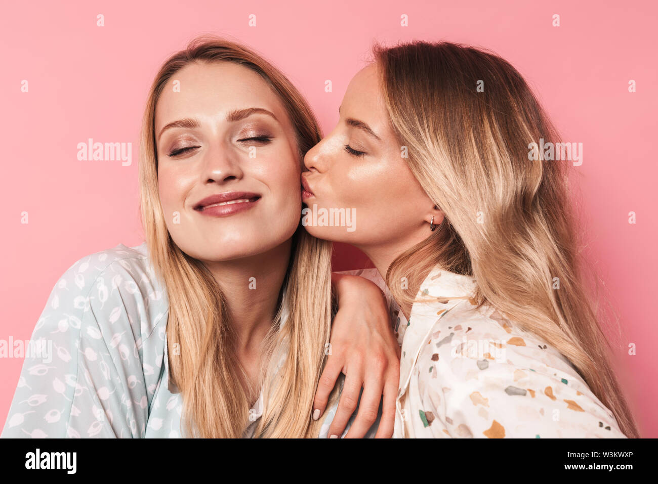 Close up of two lovely attractive blonde girls wearing summer outfit standing isolated over pink background, kissing on a cheek Stock Photo