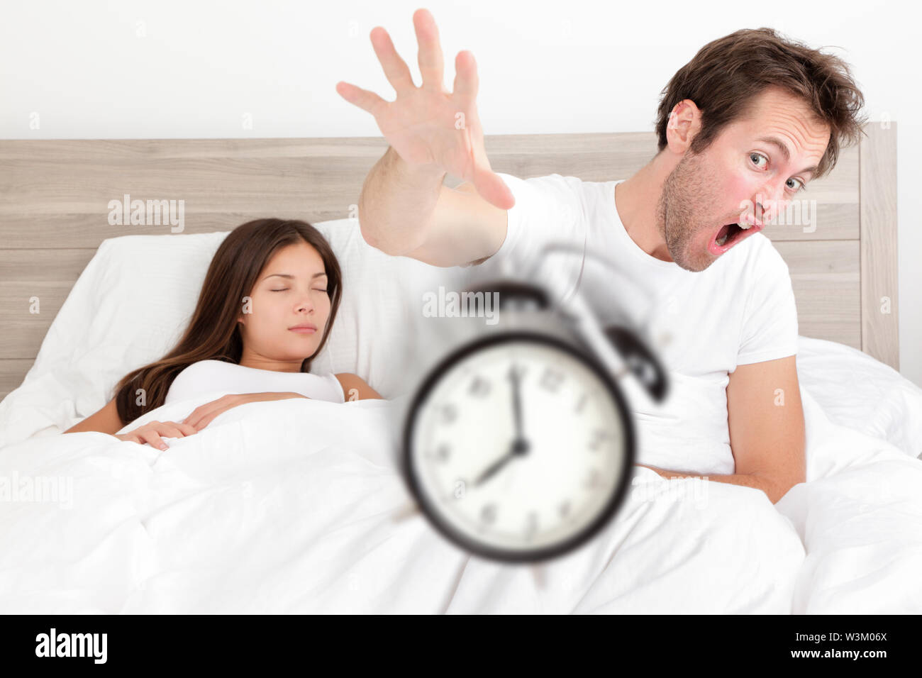 Wake up - couple waking up early throwing alarm clock. Funny bed concept with young interracial couple waking up late. Man throwing alarm clock, and woman sleeping. Asian female, Caucasian male models Stock Photo