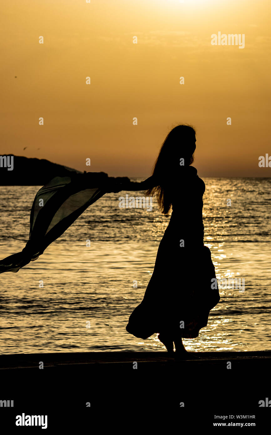 Beautiful female silhouette of unrecognizable slim tall woman with silk scarf at sunset in front of golden Ionian Sea water. Colorful dusk as seen from Ksamil, Albania, late spring Stock Photo