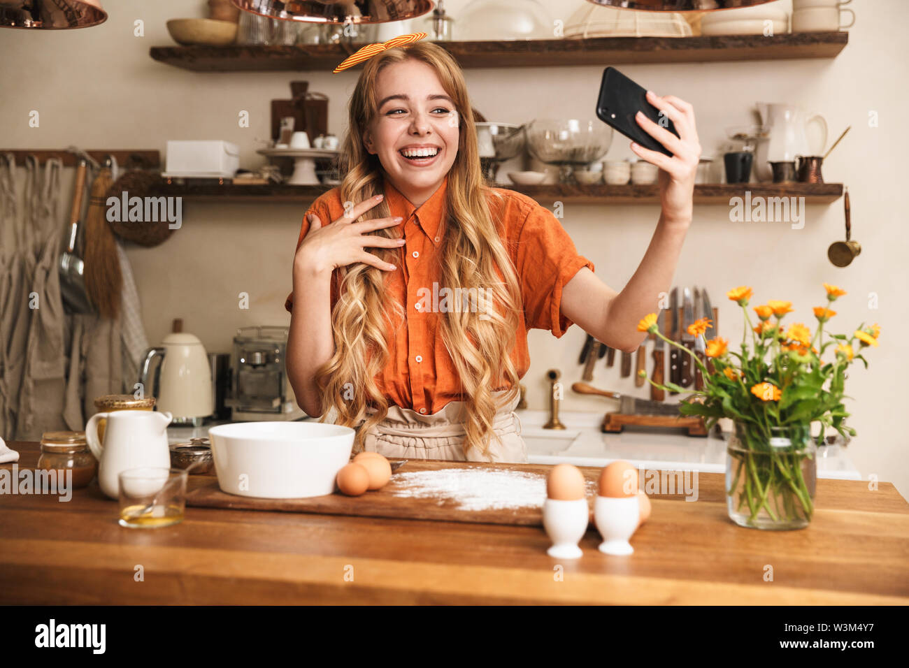 Image of a cheery young emotional blonde girl chef cooking at the kitchen take a selfie by mobile phone. Stock Photo