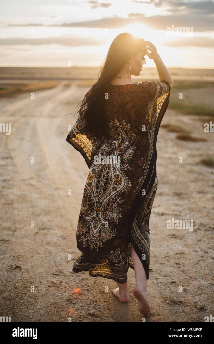 Woman wearing patterned dress walking on dirt road at sunset Stock Photo