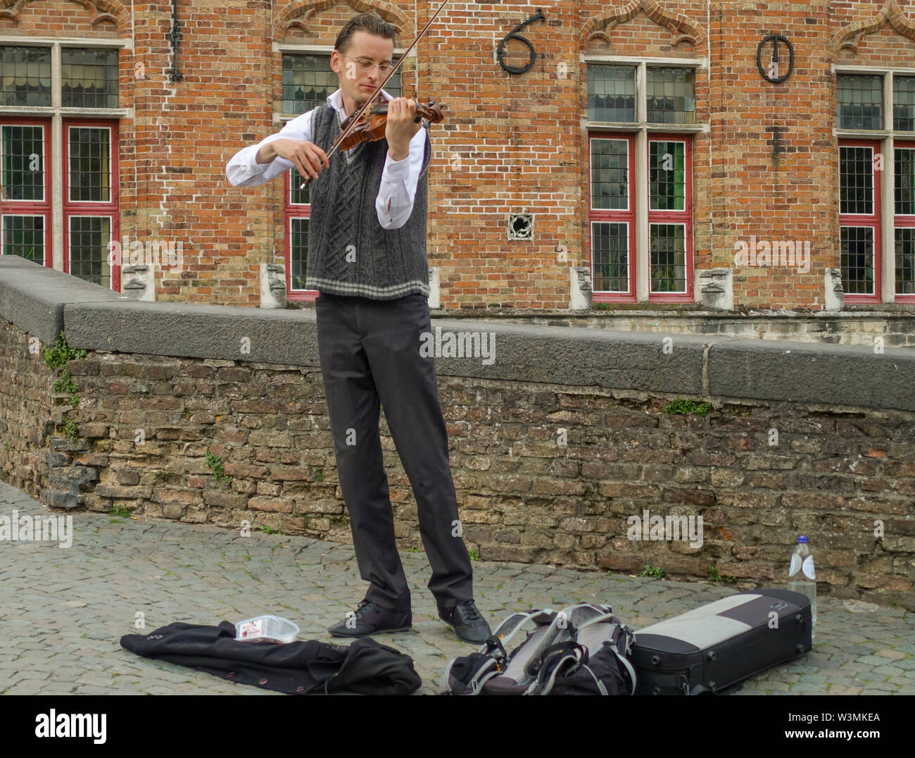 Bruges, Flanders, Belgium -  June 16, 2019: Young man plays violin as beggar for money on bridge over canal at Blinde Ezelstraat. Red brick building i Stock Photo