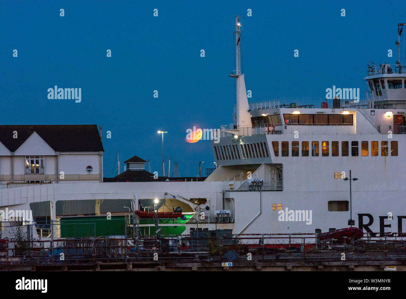 Partial lunar eclipse, Southampton, Hampshire, England, UK, 16th July 2019. A partial lunar eclipse and a hazy sky on the horizon turn the moon orange red during evening moonrise over the south coast. In the foreground, the Red Falcon Red Funnel Isle of Wight ferry has just docked in the port for disembarkation. Stock Photo