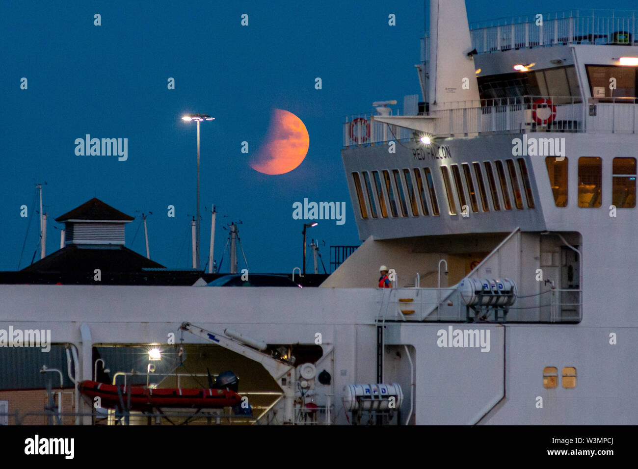 Partial lunar eclipse, Southampton, Hampshire, England, UK, 16th July 2019. A partial lunar eclipse and a hazy sky on the horizon turn the moon orange red during evening moonrise over the south coast. In the foreground, the Red Falcon Red Funnel Isle of Wight ferry has just docked in the port for disembarkation. Stock Photo