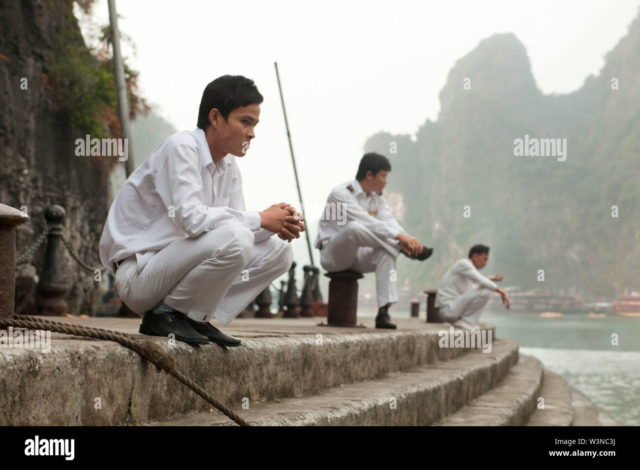 3 sailors who work as crew members on vacation tourist boats wait at dock with the lime stone towers of Ha Long Bay behind them Stock Photo
