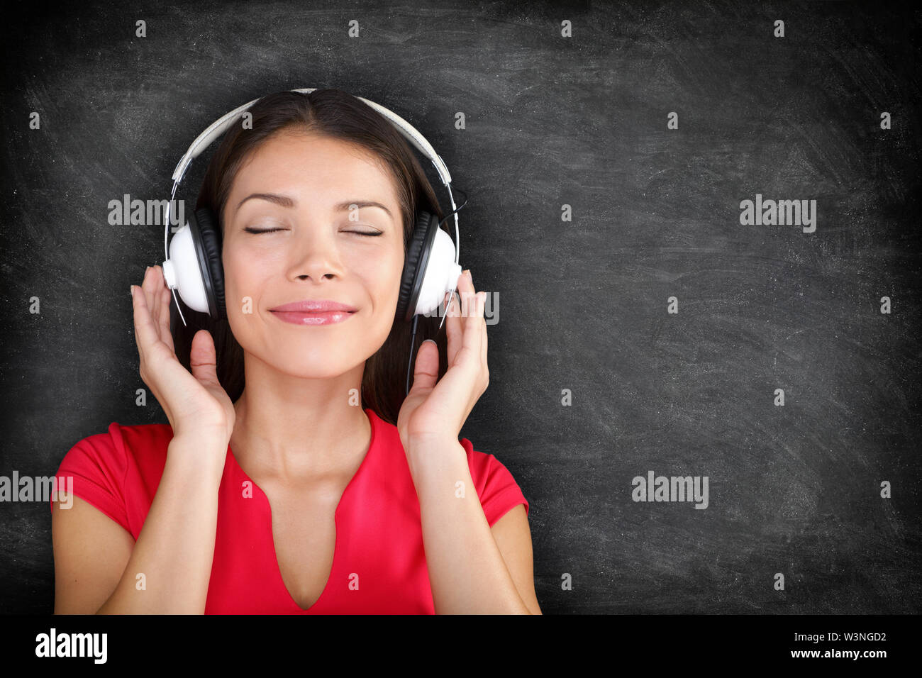 Music in headphones. Beautiful young Asian woman with her eyes closed in bliss listening to music wearing a set of headphones and standing against black blackboard background with copyspace Stock Photo