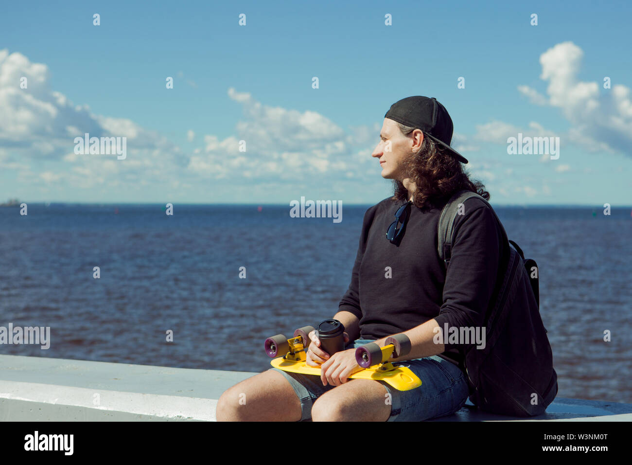 A young man with long hair in a baseball cap, denim shorts, sweatshirt and  black glasses is sitting with a yellow skate on the city's quay. Copy space  Stock Photo - Alamy