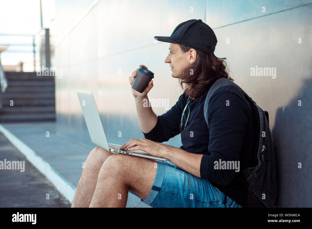 Young man with long hair and baseball cap sits on yellow skateboard on the  asphalt, leaning his back on gray granite wall, and and drinks coffee Stock  Photo - Alamy