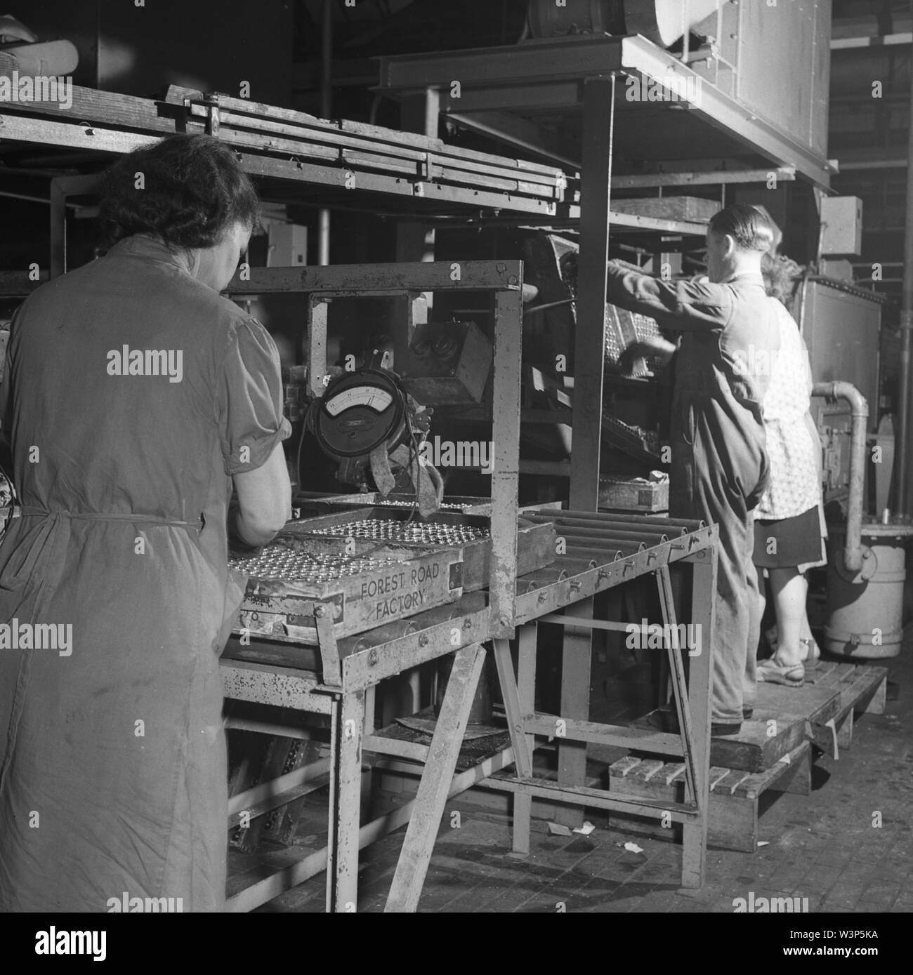 1950s, historical, male and female workers in overalls at the Ever Ready battery  factory, Forest Road, Walthamstow, London, England, UK. One female worker  is checking the meter reading of the batteries Stock