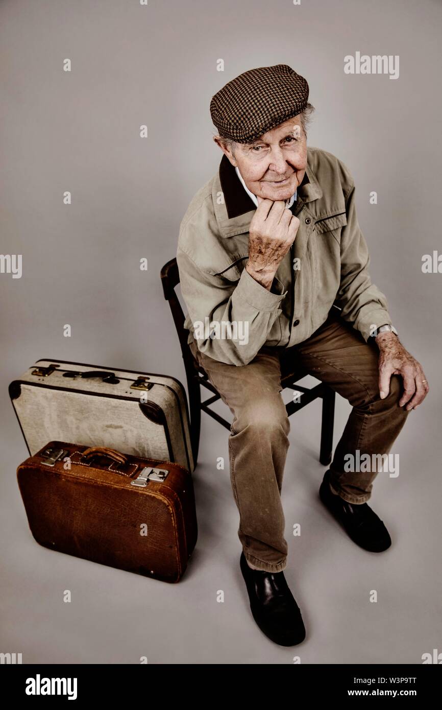Senior sits with old suitcase on a chair, symbolic picture leaving, longing, studio shot, Germany Stock Photo