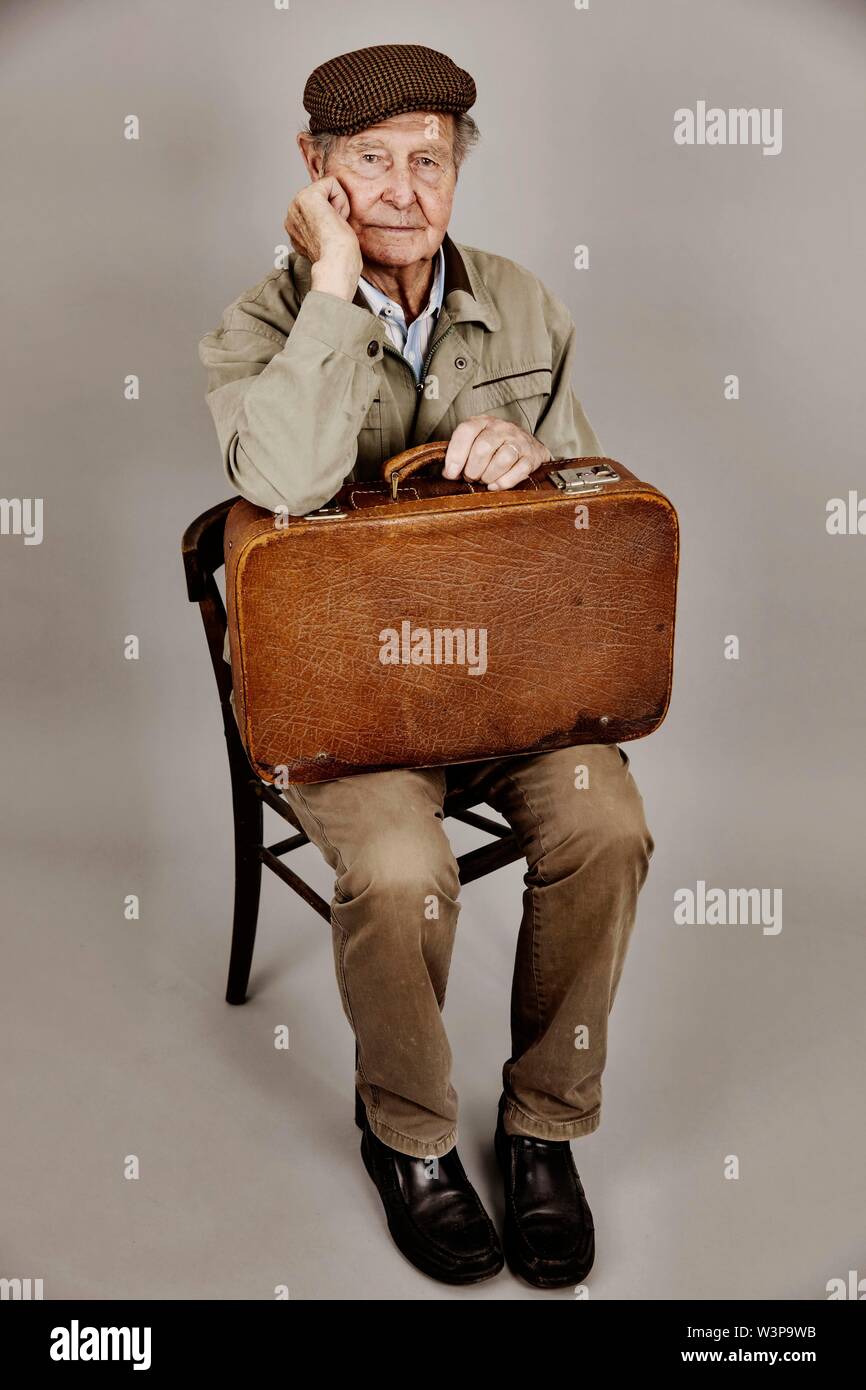 Senior sits with old suitcase on a chair, symbolic picture leaving, longing, studio shot, Germany Stock Photo