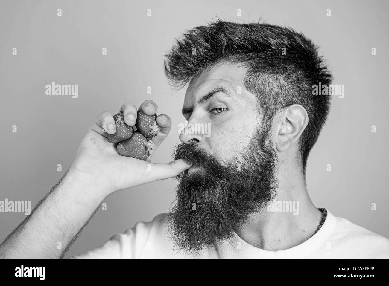 Hipster bearded holds strawberries fist as juice bottle. Man drinks strawberry juice suck thumb as drink straw blue background. Man strict face enjoy fresh drink strawberry juice. Fresh juice concept. Stock Photo