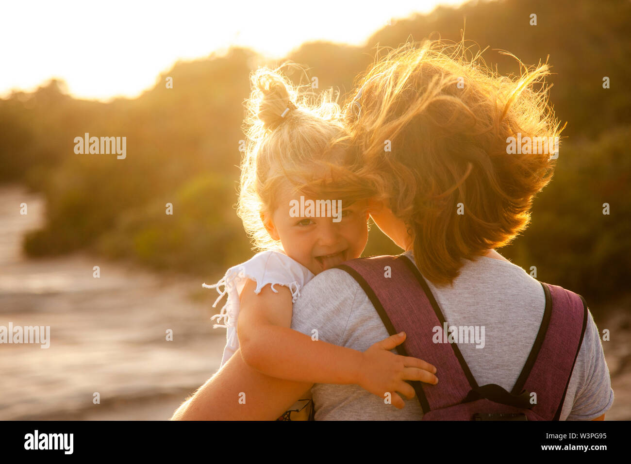portrait of a 3 year old Caucasian girl with blond hair who shows her tongue playfully. In the arms of the mother, backlit image at sunset. Stock Photo