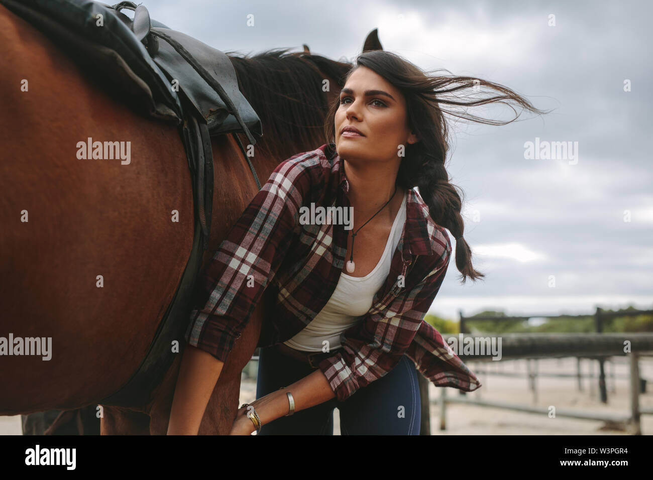 Young woman harnessing a horse at stable place. Cowgirl getting ready for horseback riding. Stock Photo